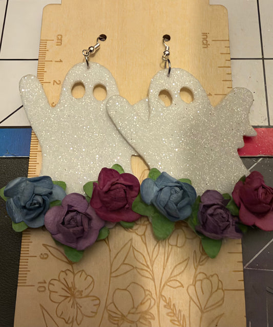 A pair of handmade ghost-shaped earrings covered in white glitter, decorated with a row of fabric roses in blue, purple, and magenta with green leaves, hanging from silver clips on a wooden ruler.