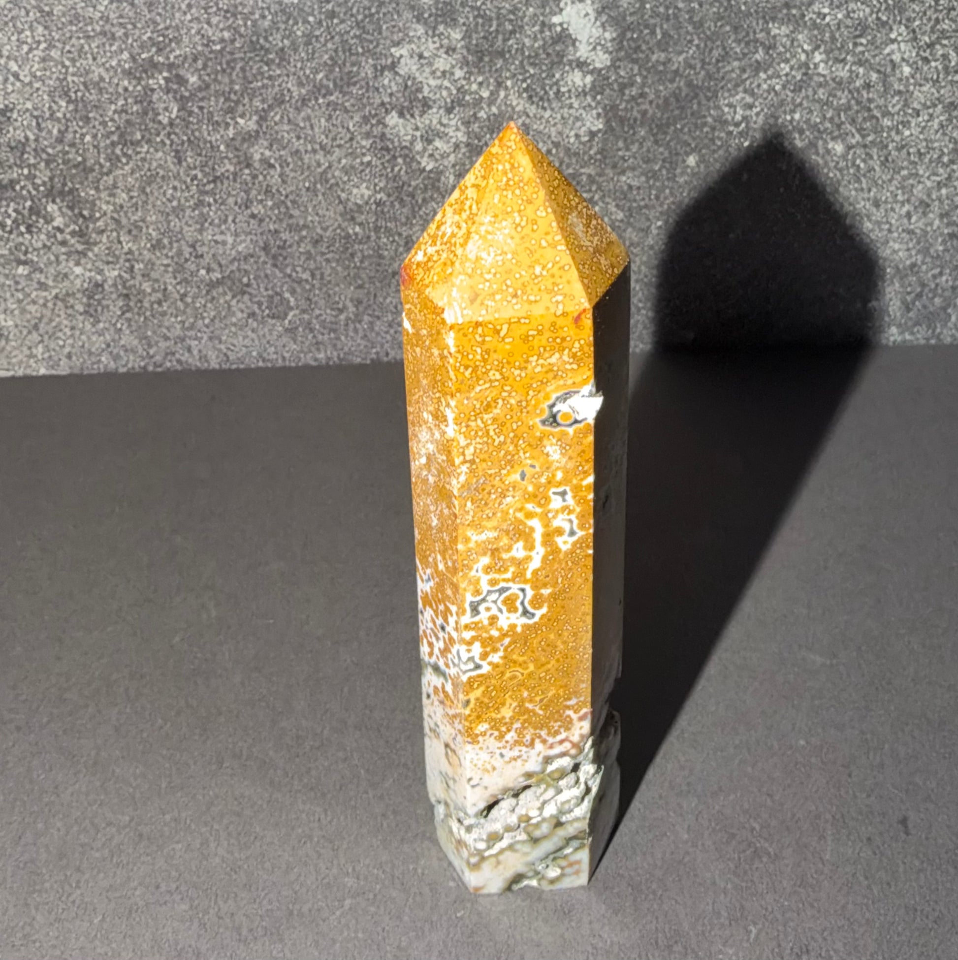 A natural golden-yellow crystal point with white and gray matrix inclusions, standing upright on a dark gray surface against a textured gray background.