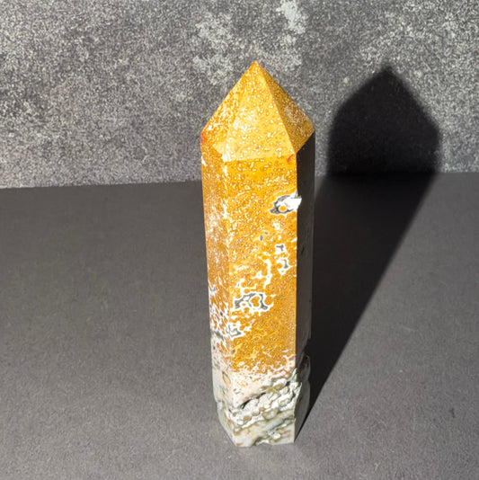 A natural golden-yellow crystal point with white and gray matrix inclusions, standing upright on a dark gray surface against a textured gray background.