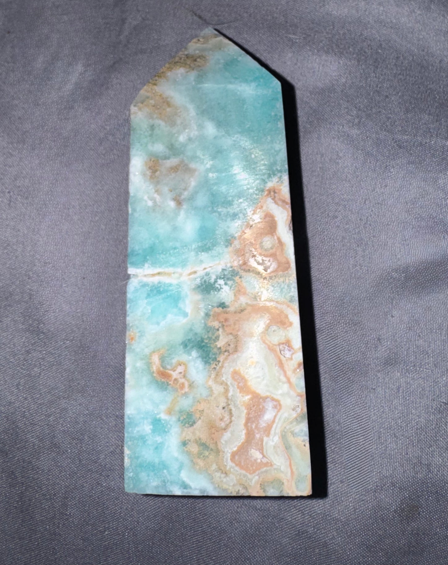 A pointed rectangular polished stone or crystal with a marbled pattern of turquoise, beige, and white, photographed upright on a dark gray textured fabric background.