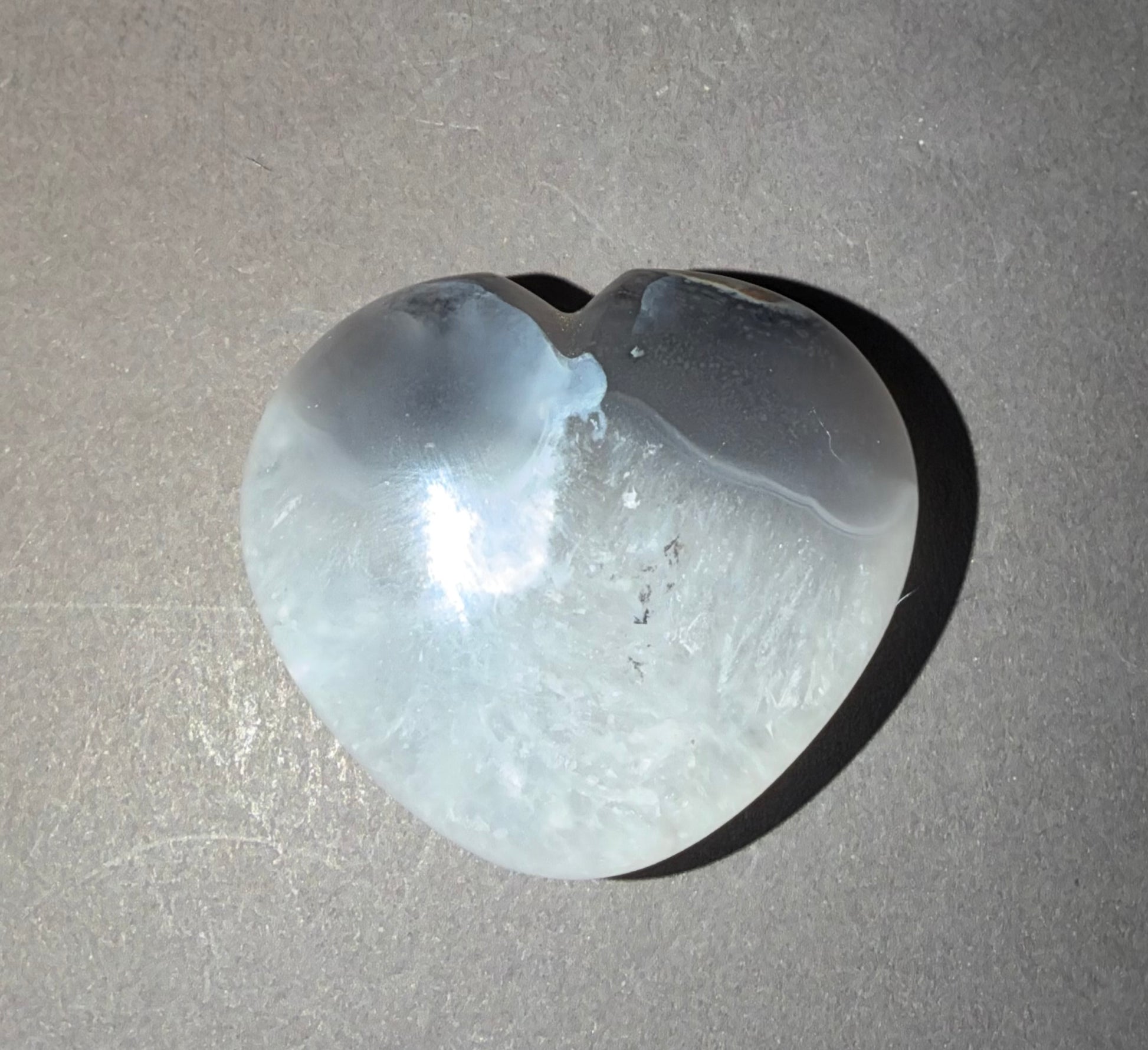 A heart-shaped translucent stone carving, likely quartz, with natural banding and inclusions, resting on a textured gray surface.