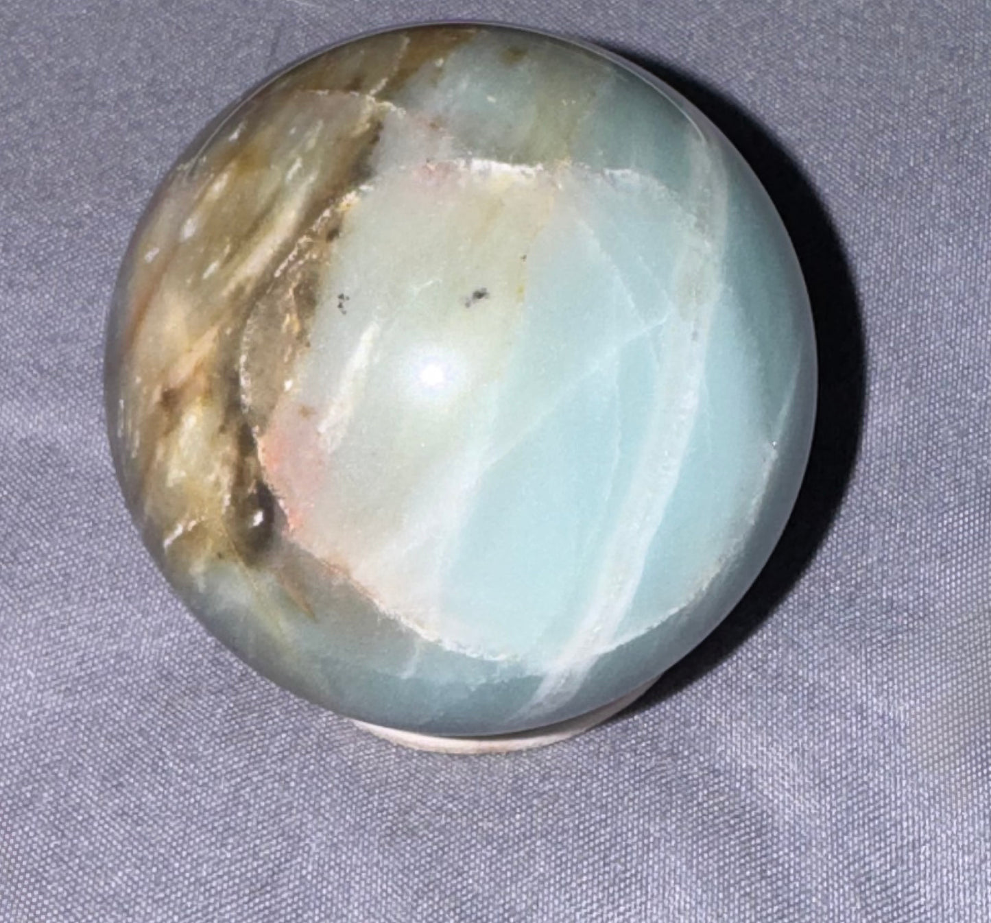 A polished round crystal sphere with natural stone patterns in light blue, white, and brown, resting on a gray textured fabric background.