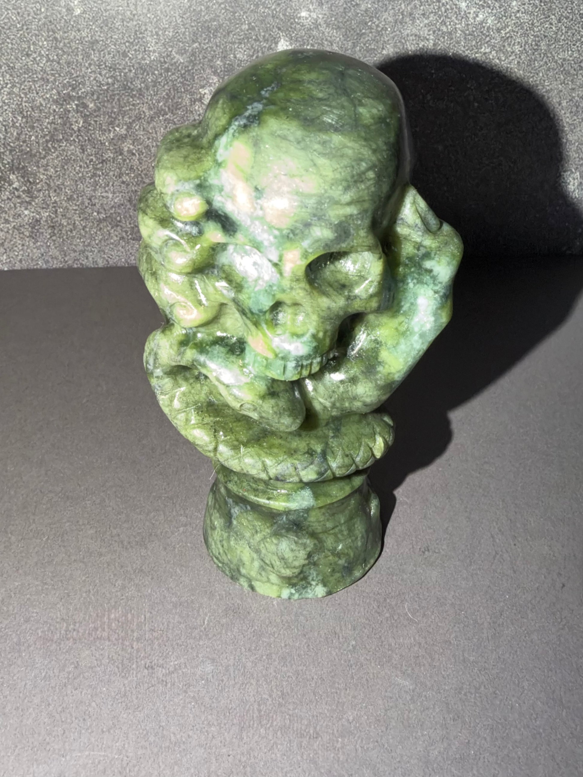 A green stone skull sculpture with carved details, standing on a matching stone pedestal against a dark gray background.