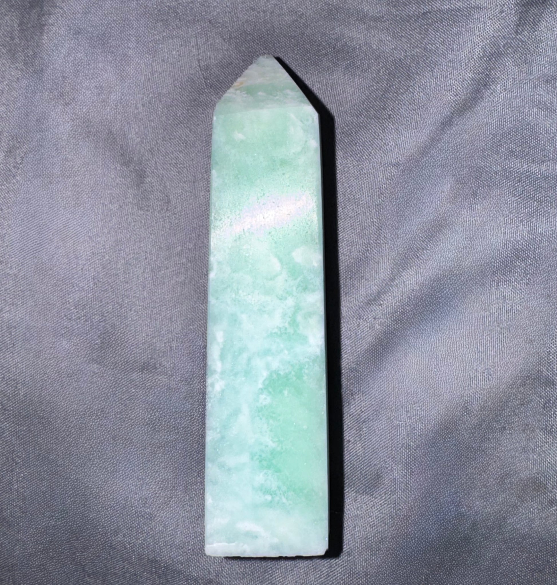A pointed green jade crystal tower or wand, showing natural light green and white marbled patterns, resting on a textured gray fabric background.