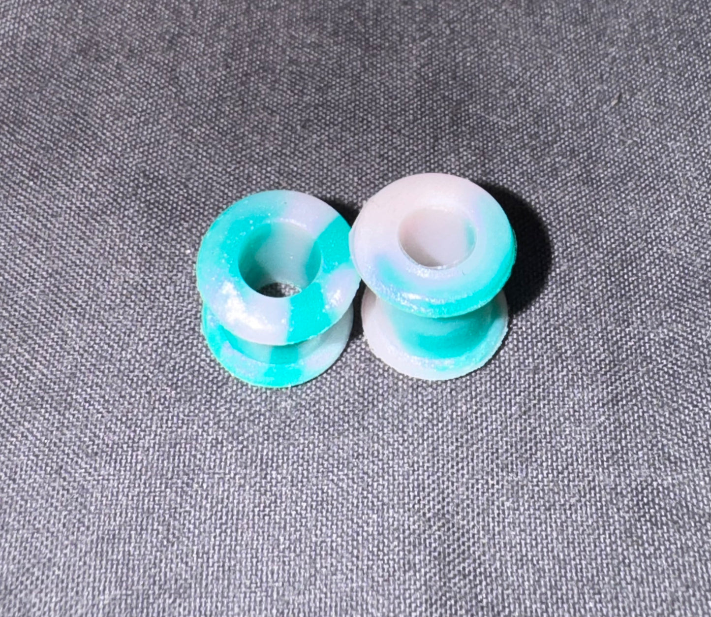 Two small round plug beads with a marbled turquoise and white resin finish, displayed on a textured gray fabric background.