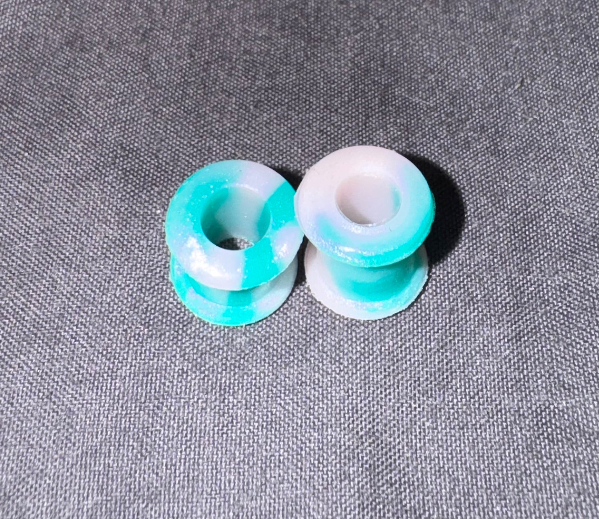 Two small round plug beads with a marbled turquoise and white resin finish, displayed on a textured gray fabric background.