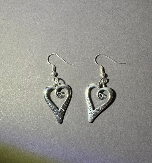 A pair of silver heart-shaped dangle earrings with decorative swirl and dot patterns, shown on a textured gray background.