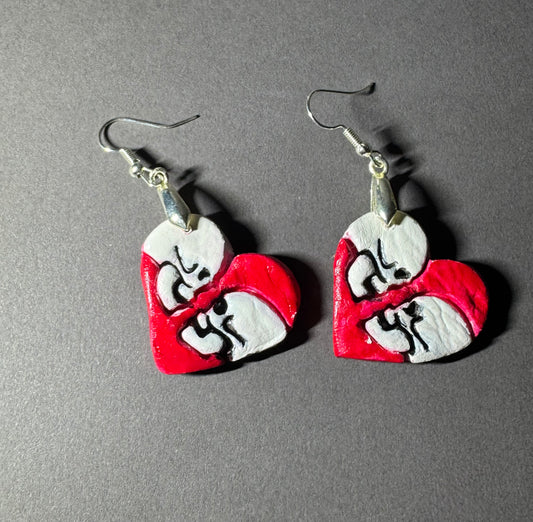 A pair of handmade heart-shaped dangle earrings featuring a split red and white design with black hand-drawn facial features, hanging from silver fishhook wires on a dark gray background.