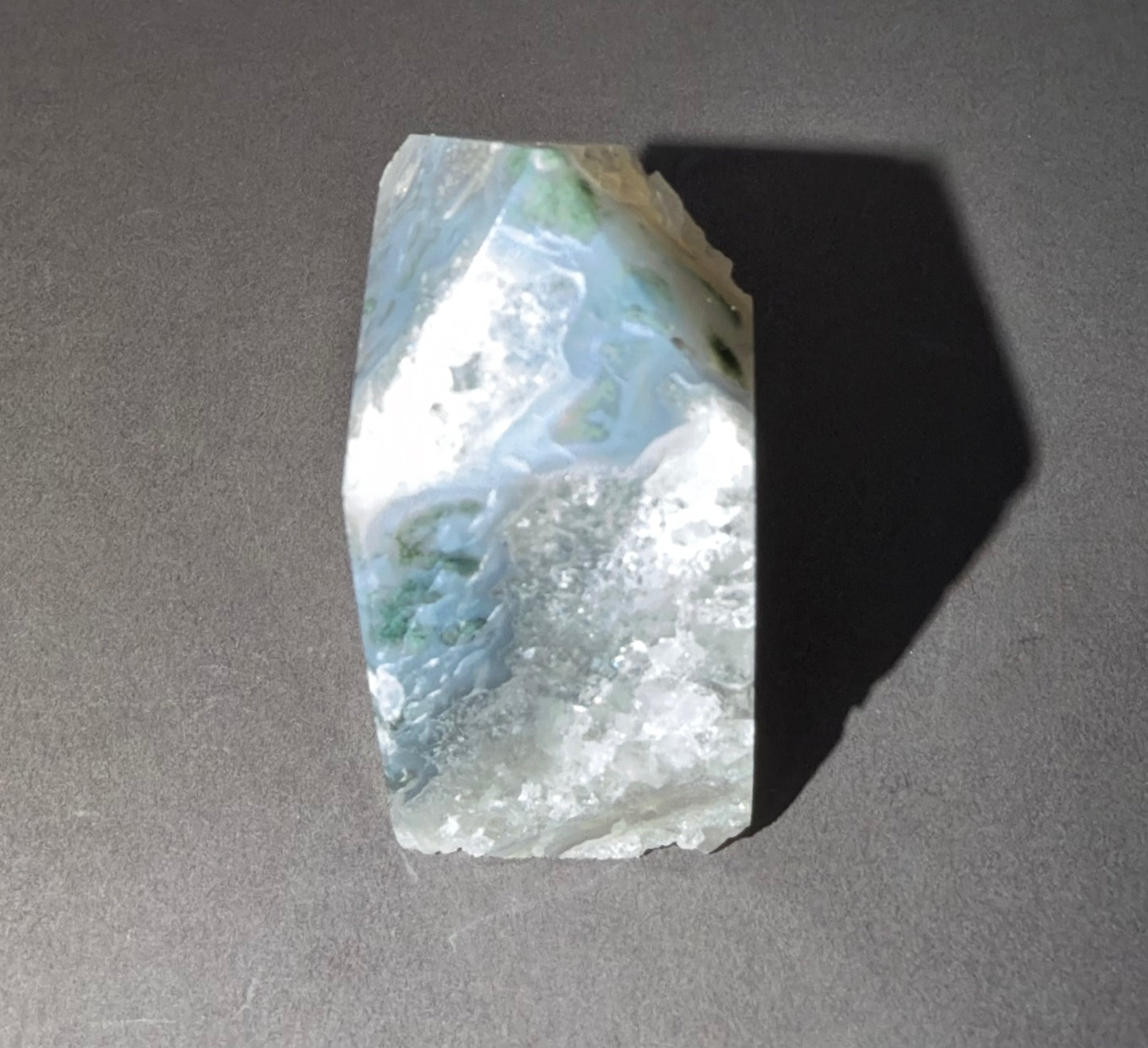 A raw, polished mineral specimen with swirling patterns of white, light blue, and green, standing upright on a dark gray surface and casting a long shadow.