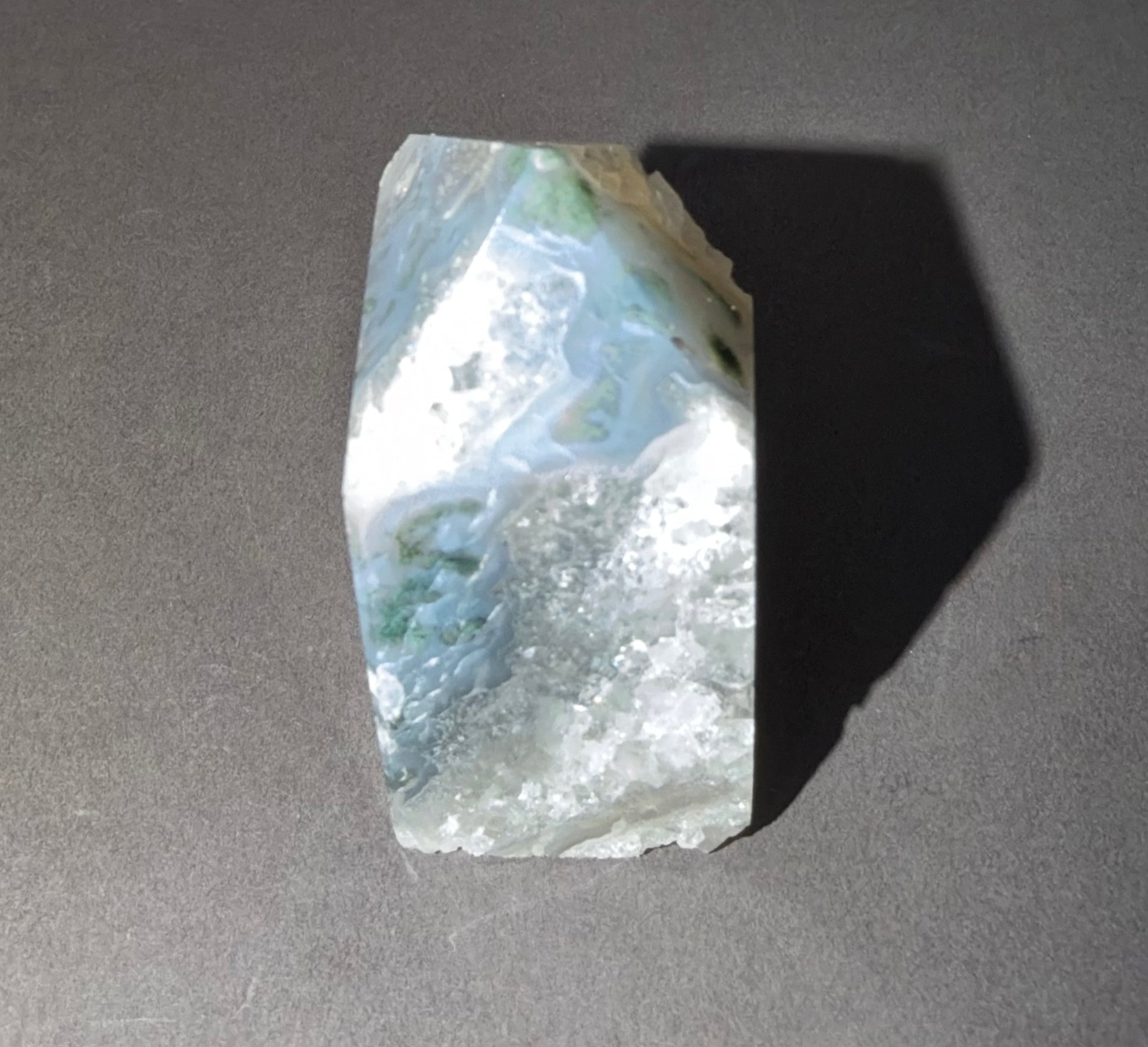 A raw, polished mineral specimen with swirling patterns of white, light blue, and green, standing upright on a dark gray surface and casting a long shadow.