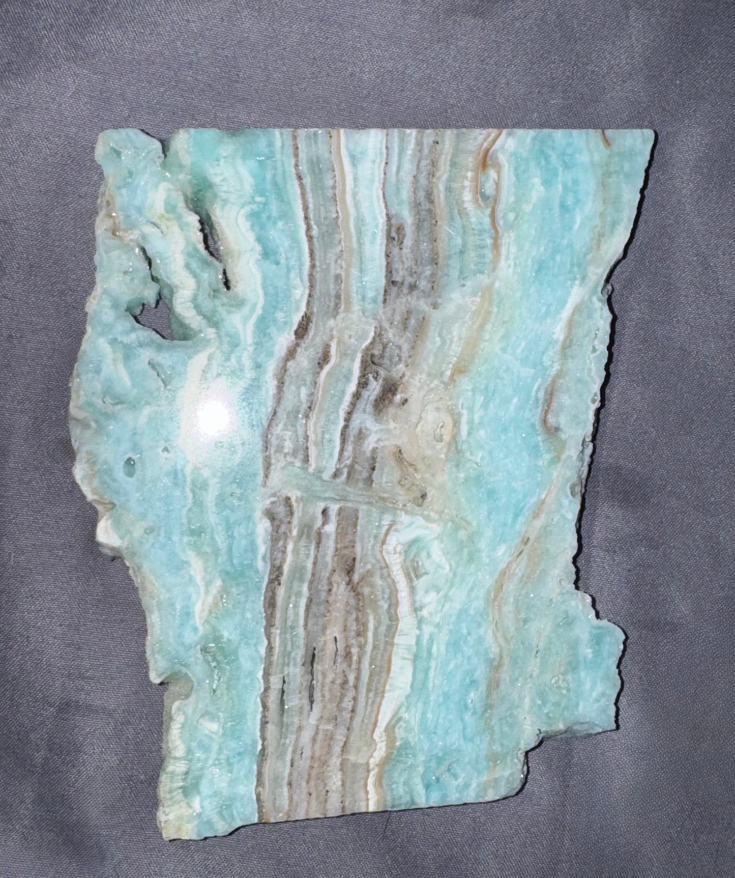 A raw polished stone slab with natural irregular edges, featuring light blue, white, and tan banded patterns, displayed on a dark gray textured background.