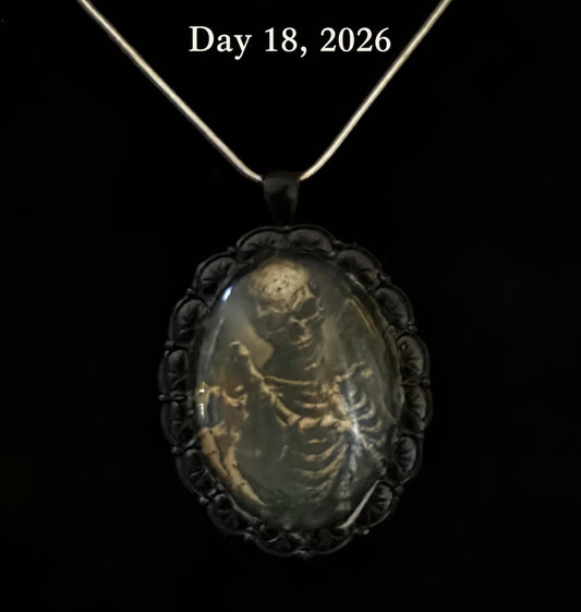 A gothic-style pendant necklace featuring a black ornate frame and a translucent oval centerpiece with a detailed skeleton illustration, suspended from a silver chain against a black background with the text 'Day 18, 2026' at the top.