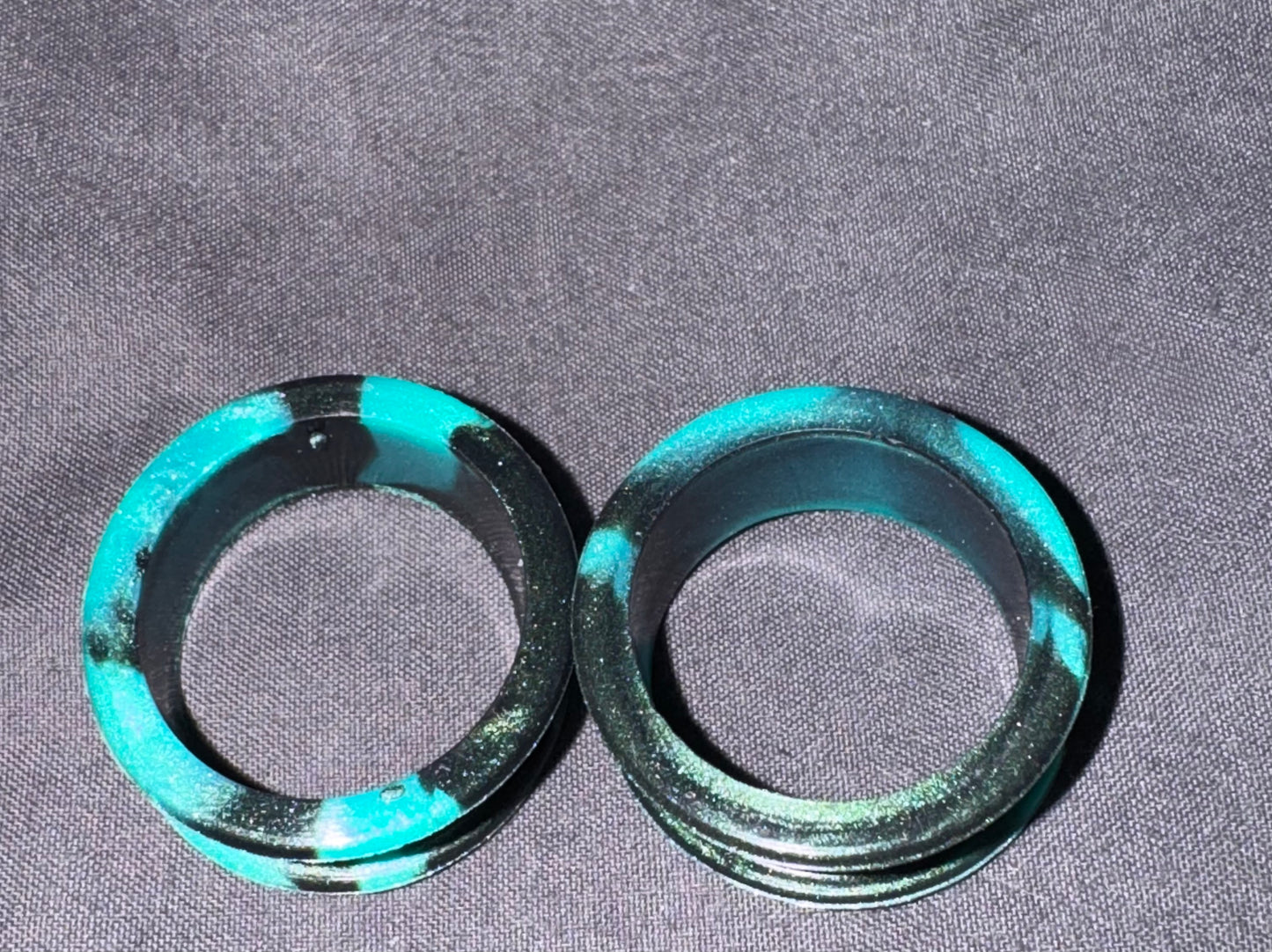 Two circular black rings with vibrant turquoise green marbled accents, displayed on a textured gray fabric background.