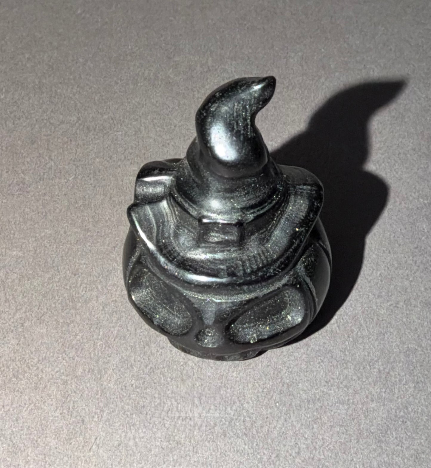 A small black carved figurine of a wizard wearing a pointed hat, photographed on a neutral gray background with a sharp shadow cast to the right.