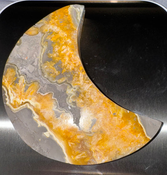 A crescent-shaped decorative stone slab with a marbled pattern of orange, yellow, gray, and white, displayed on a dark metallic surface.