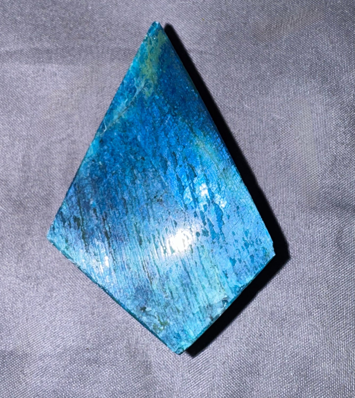 A triangular blue gemstone or polished crystal with a textured, polished surface, resting on a gray fabric background.