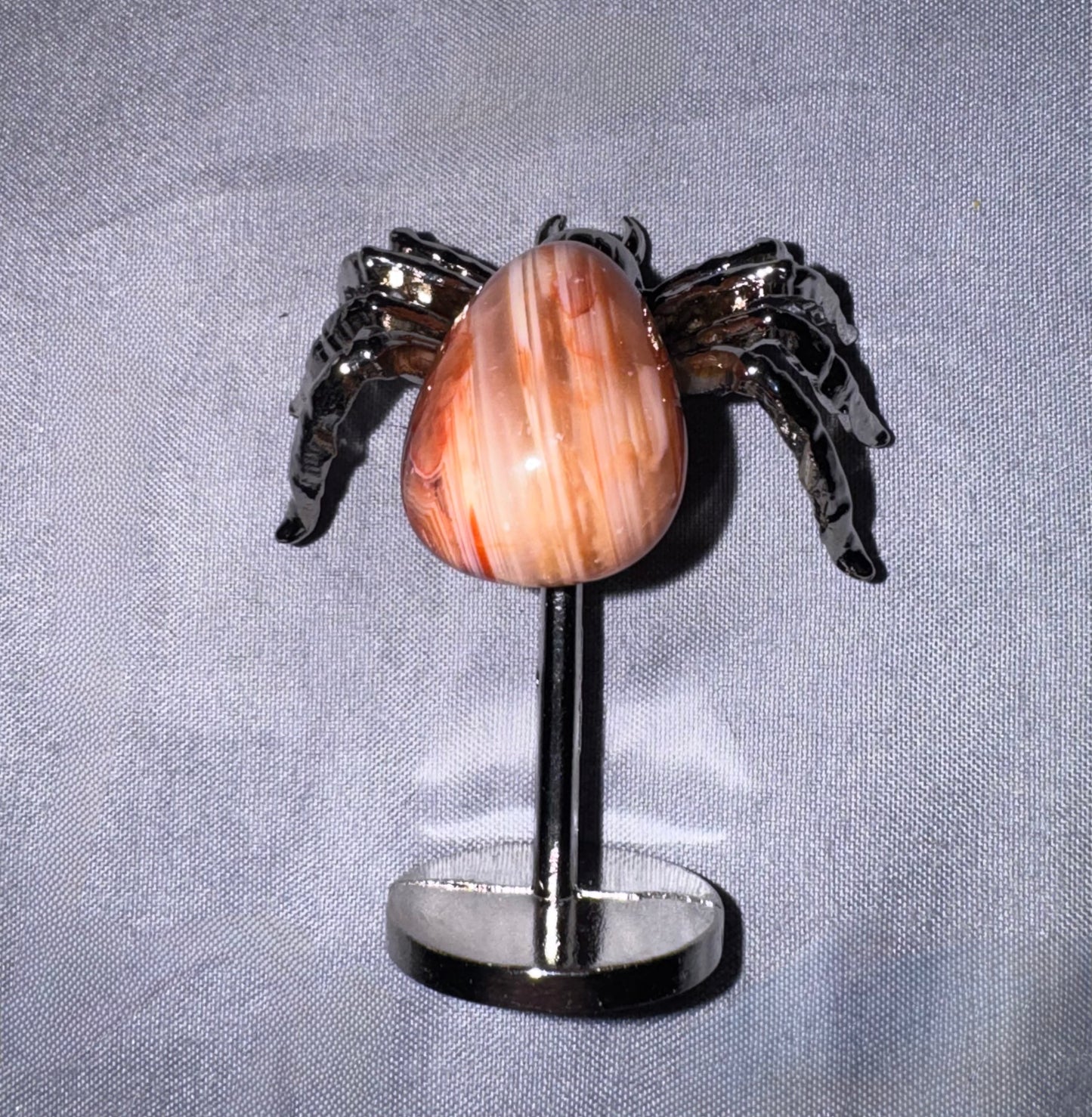 A spider with a polished teardrop-shaped peach and cream banded stone center and dark metallic spider legs, mounted on a black display stand against a light gray textured fabric background.