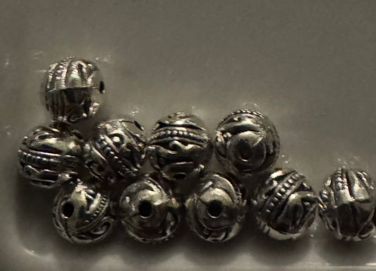 A group of small metallic silver beads with intricate cutout and textured patterns, arranged on a plain light gray surface.