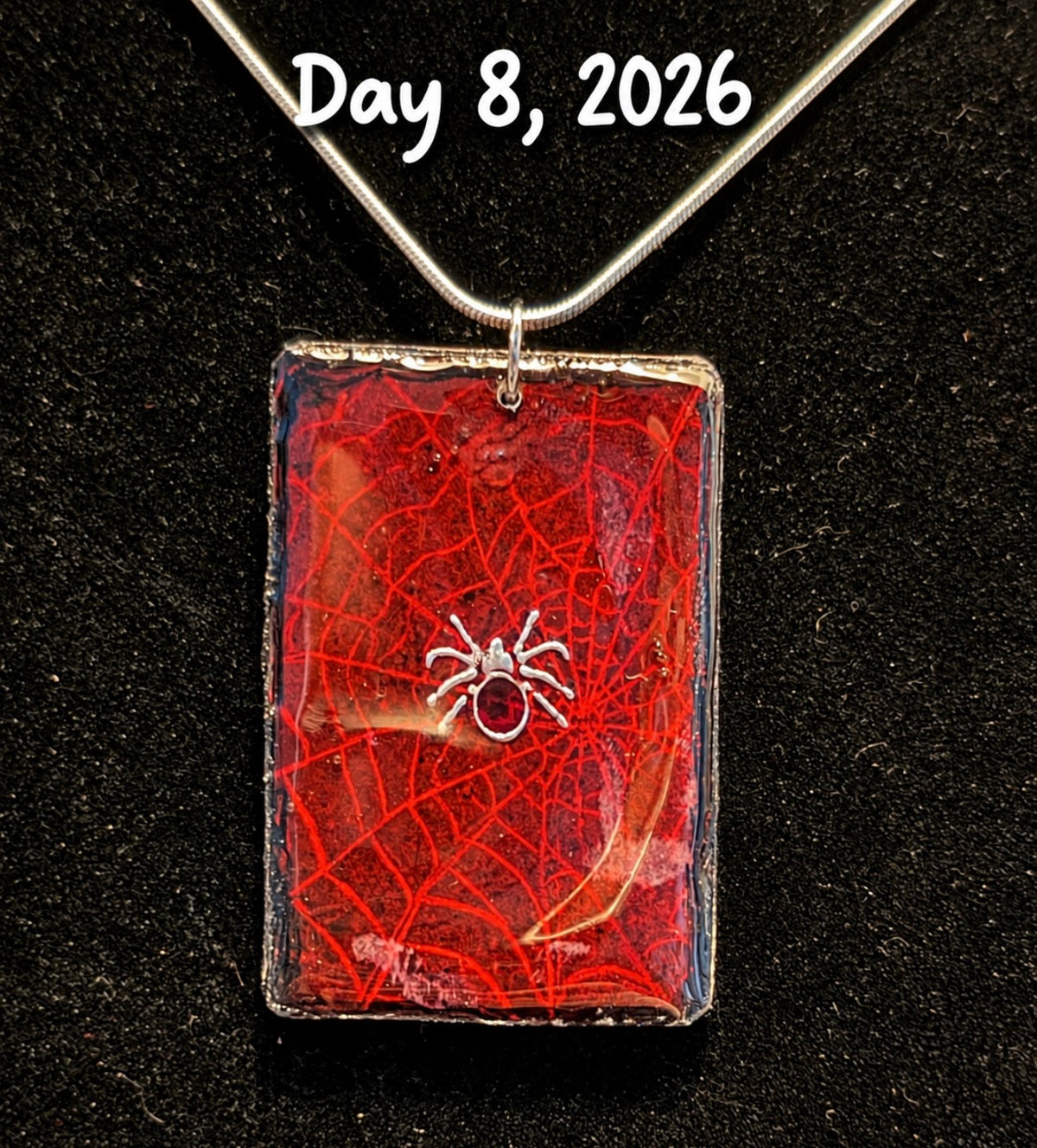 A handmade rectangular pendant necklace featuring a red resin background with a web-like pattern and a small silver spider in the center, suspended from a silver chain on a black background. White text at the top reads 'Day 8, 2026'.