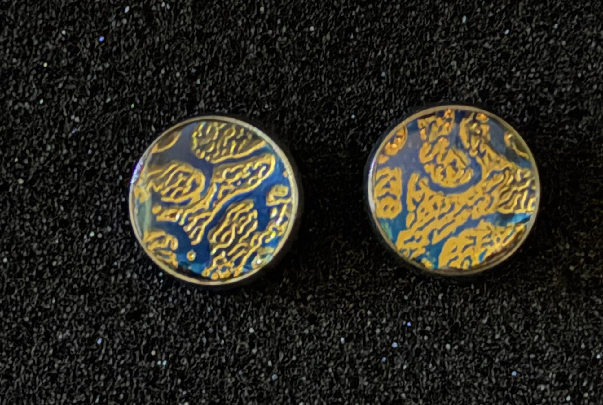 A pair of round stud earrings with a textured gold and blue abstract pattern, displayed on a black textured background.