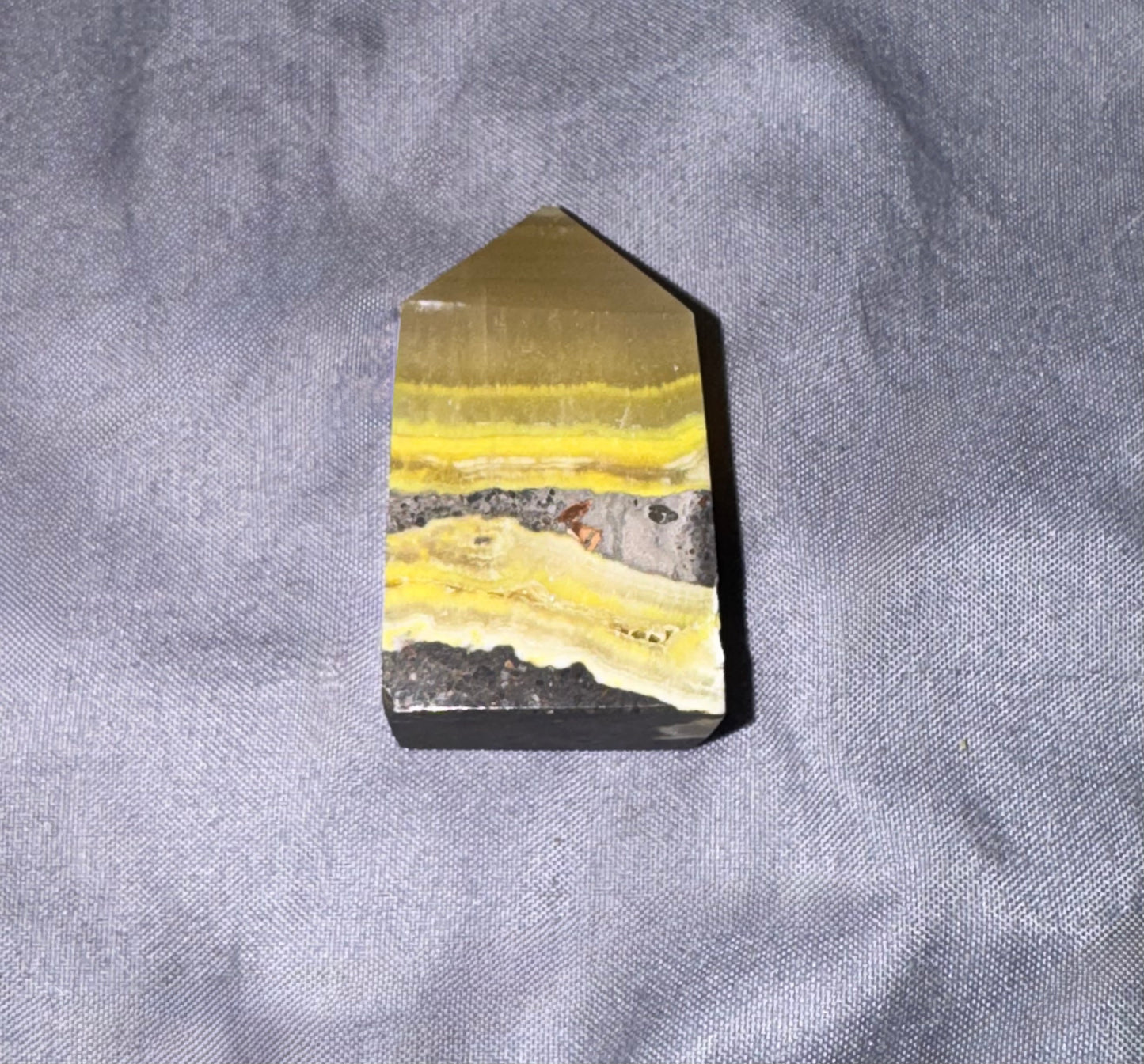 A pointed crystal tower or mineral specimen with layered yellow, gray, and black bands, photographed on a textured gray fabric background.