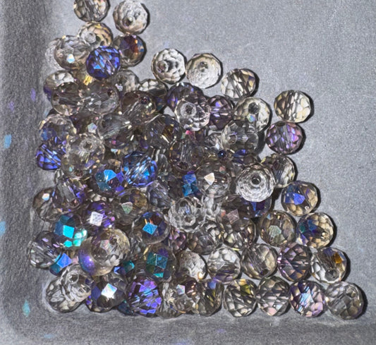 A pile of faceted round craft beads with a smoky gray base and iridescent blue, purple, and gold AB finish, resting on a textured gray background.