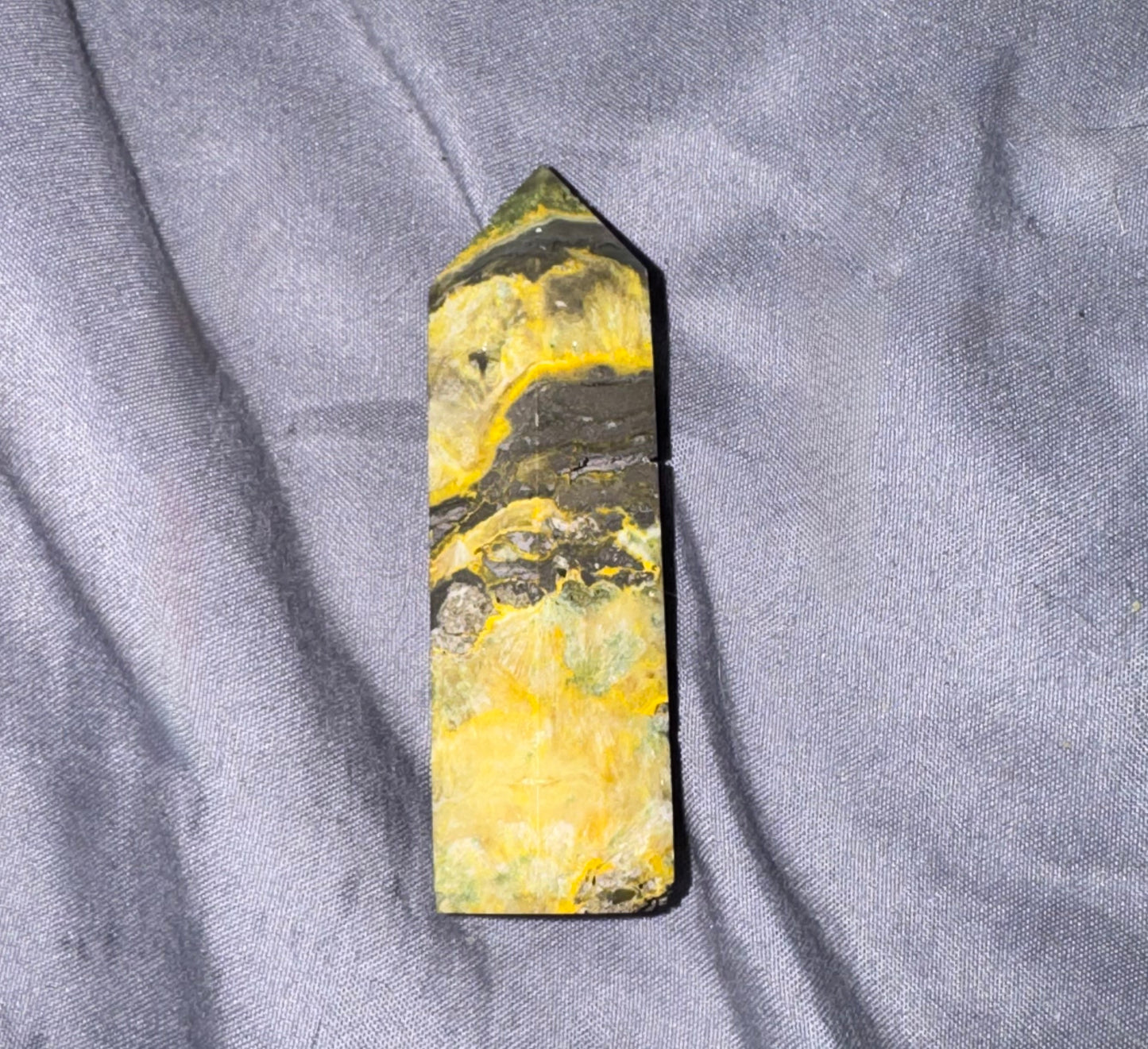 A polished natural crystal point with marbled yellow, green, and dark gray/black patterns, resting on a textured gray fabric background.
