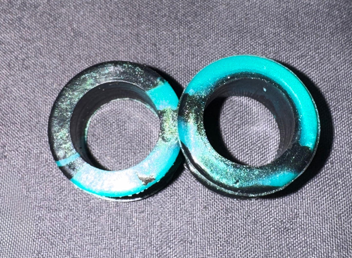 Two circular black rubber or plastic rings with bright turquoise blue painted sections, placed on a gray textured fabric background.