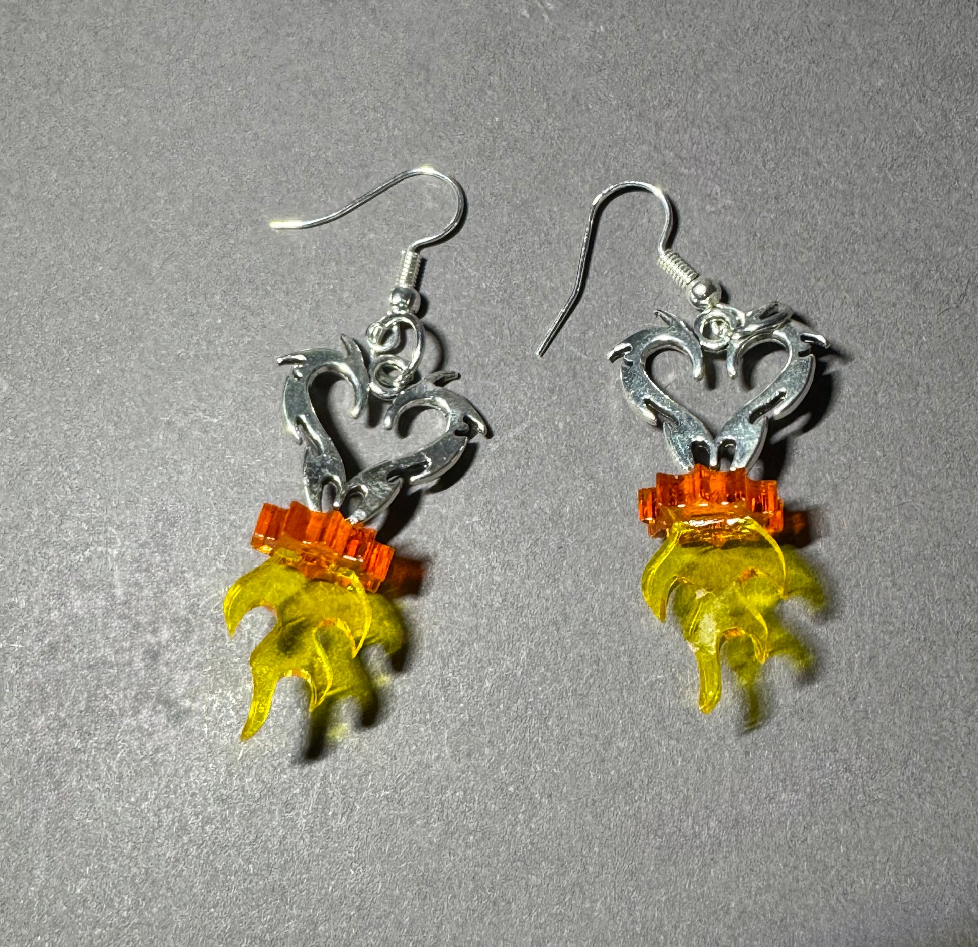 A pair of handmade dangle earrings featuring silver-toned metal filigree, orange beads, and translucent yellow flame-shaped charms, displayed on a textured gray background.