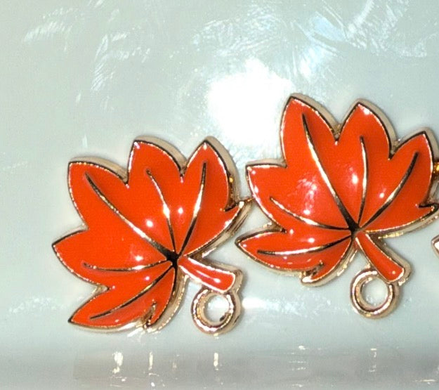 A pair of orange maple leaf charms with gold-tone metal borders and small hanging loops, shown on a glossy white surface.
