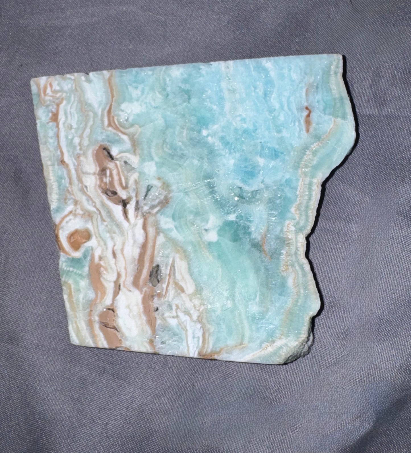 A raw, polished stone slab with natural jagged edges, featuring marbled patterns of light blue, white, and tan, displayed on a textured gray fabric background.