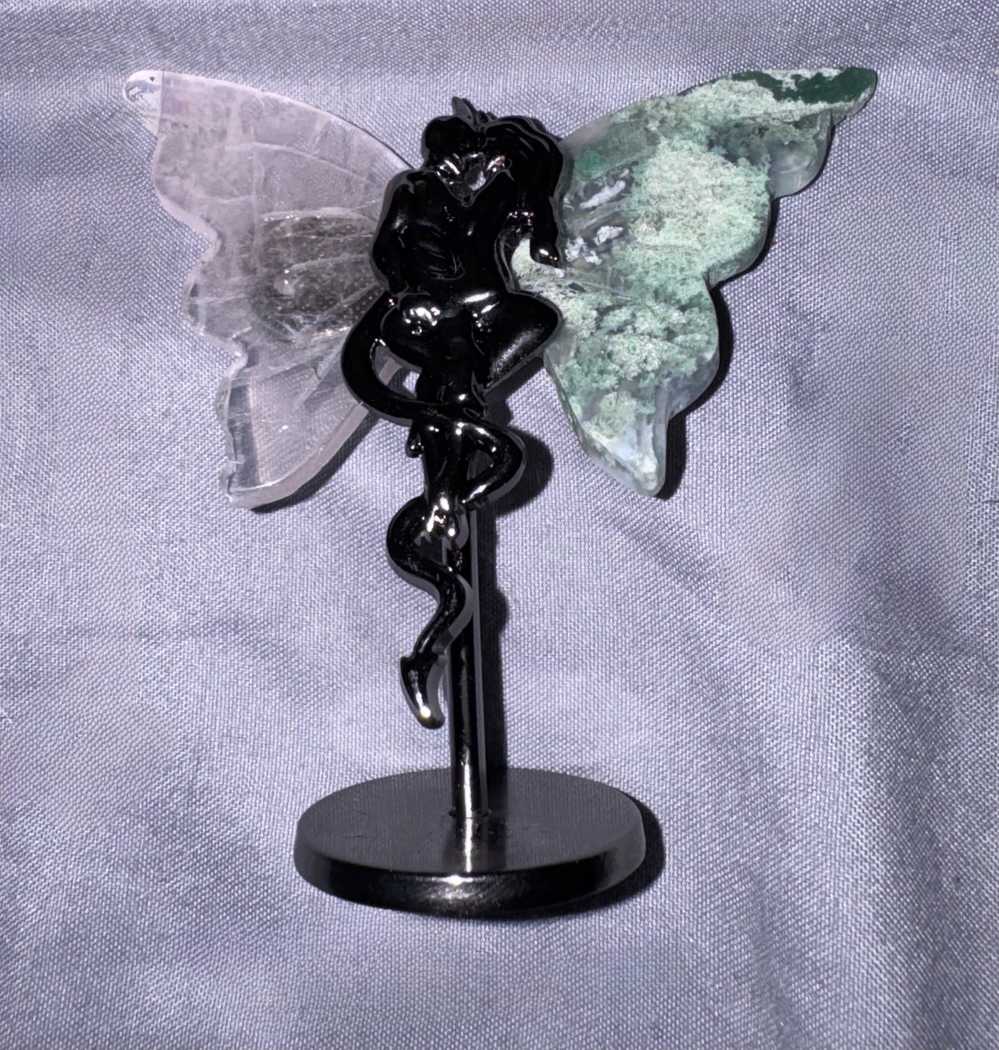 A  figurine with a glossy black body and a translucent clear wing, paired with a textured green wing, mounted on a black circular stand against a gray fabric background.