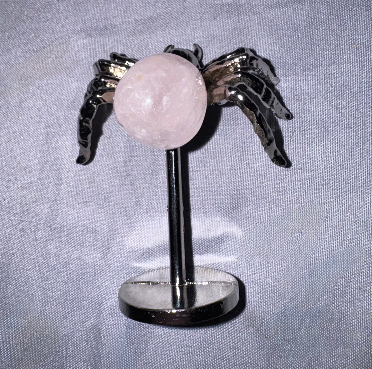 A small decorative object featuring a pale pink round stone or crystal on a black stand, with two dark metallic wing-like shapes extending from the top, photographed against a light fabric background.