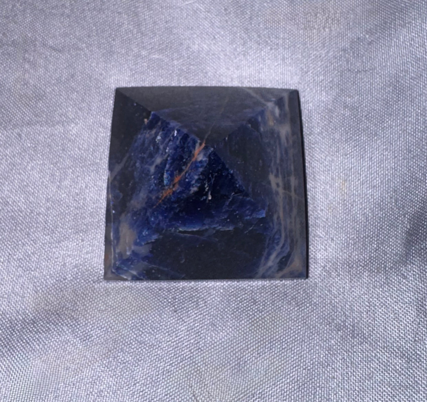A polished dark blue and black stone pyramid cabochon with natural inclusions, displayed on a textured gray fabric background.