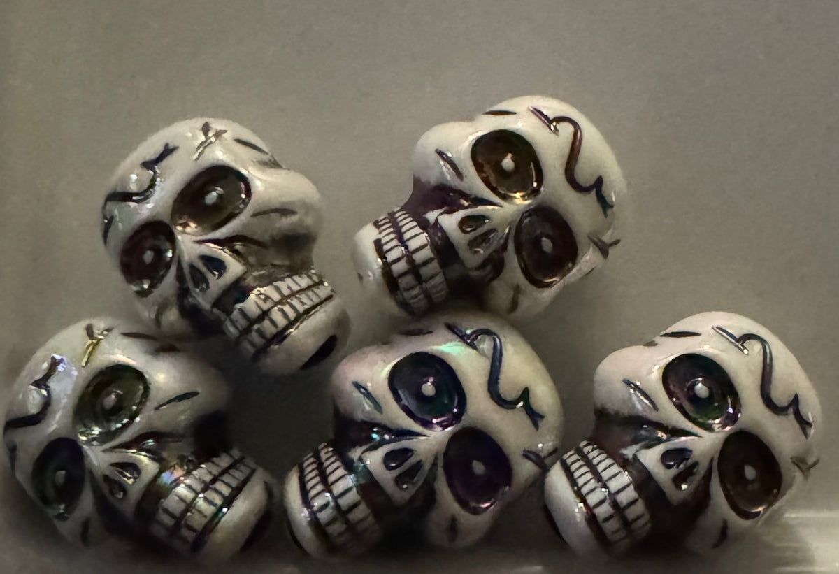 Five white sugar skull beads with black painted details and iridescent purple, green, and gold accents, arranged on a neutral gray background.
