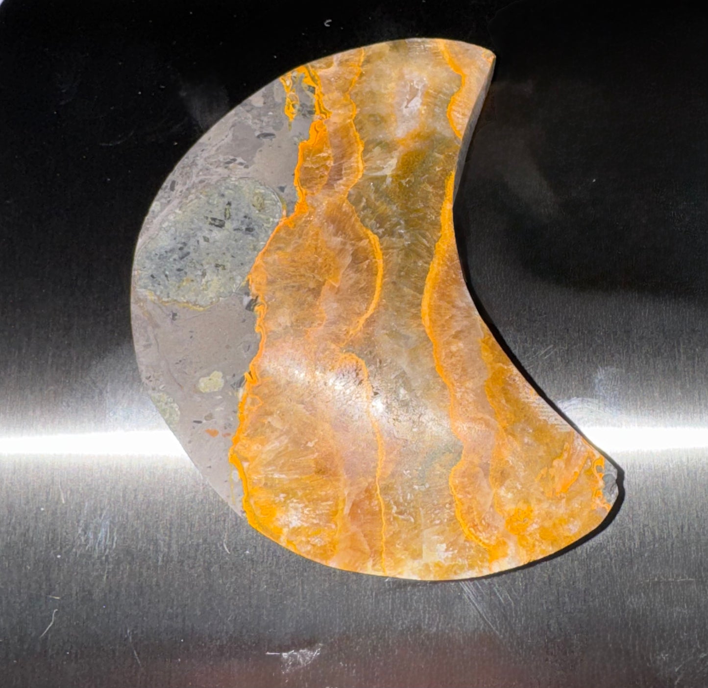 A crescent-shaped polished stone beauty tool with vibrant orange and yellow banded patterns over a gray base, resting on a brushed metal surface.