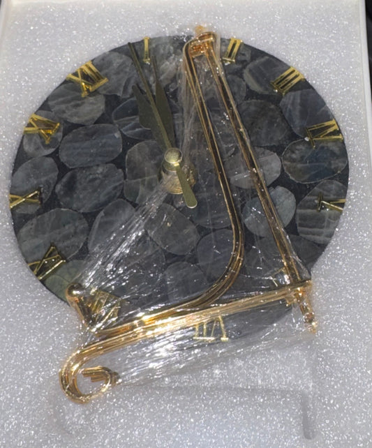 A round wall clock with a dark gray stone-pattern face and gold Roman numerals, wrapped in clear plastic and resting on white foam packaging.