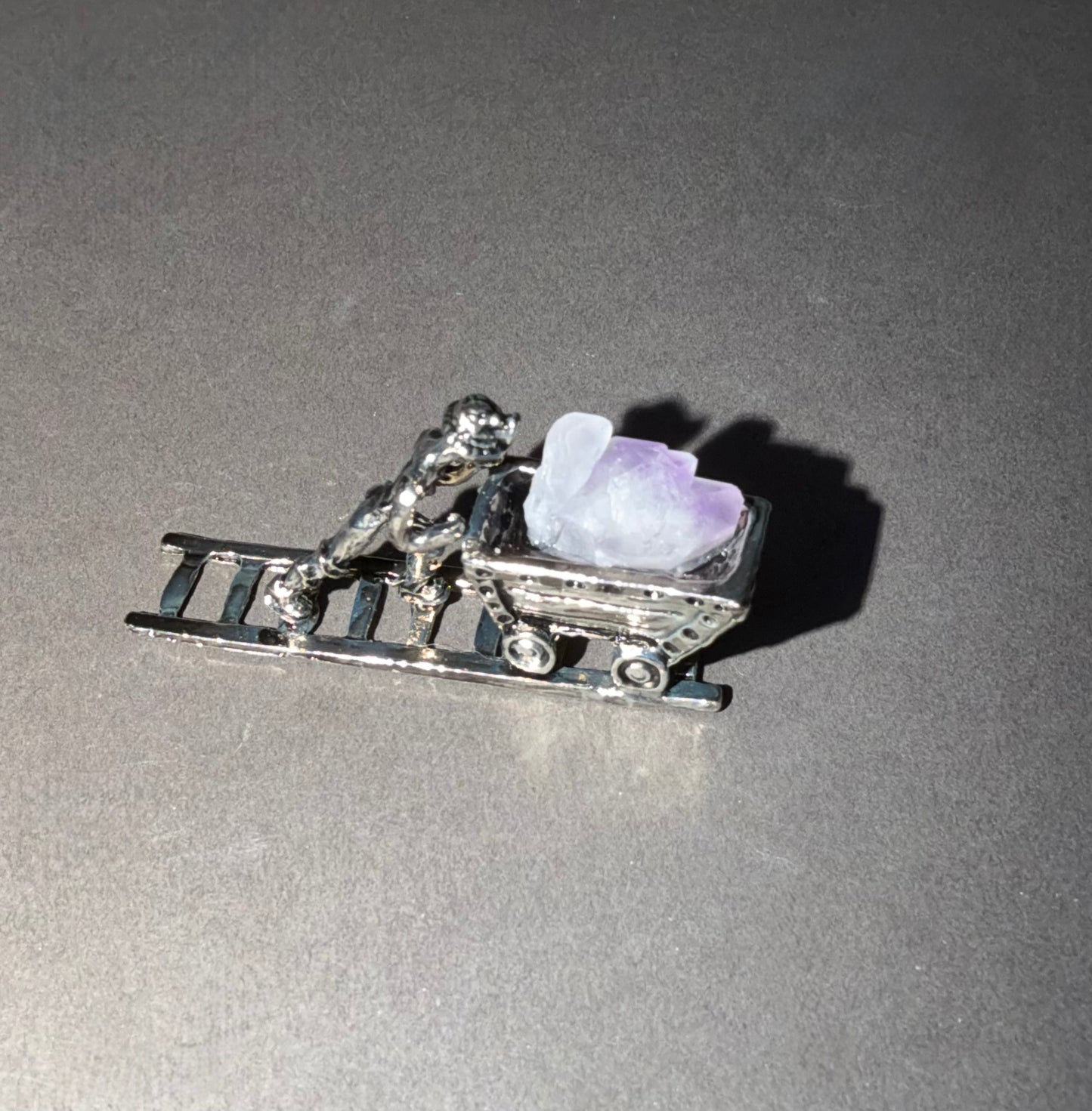 A silver-toned miniature charm depicting a person pushing a sled with a light purple crystal, shown on a gray background.