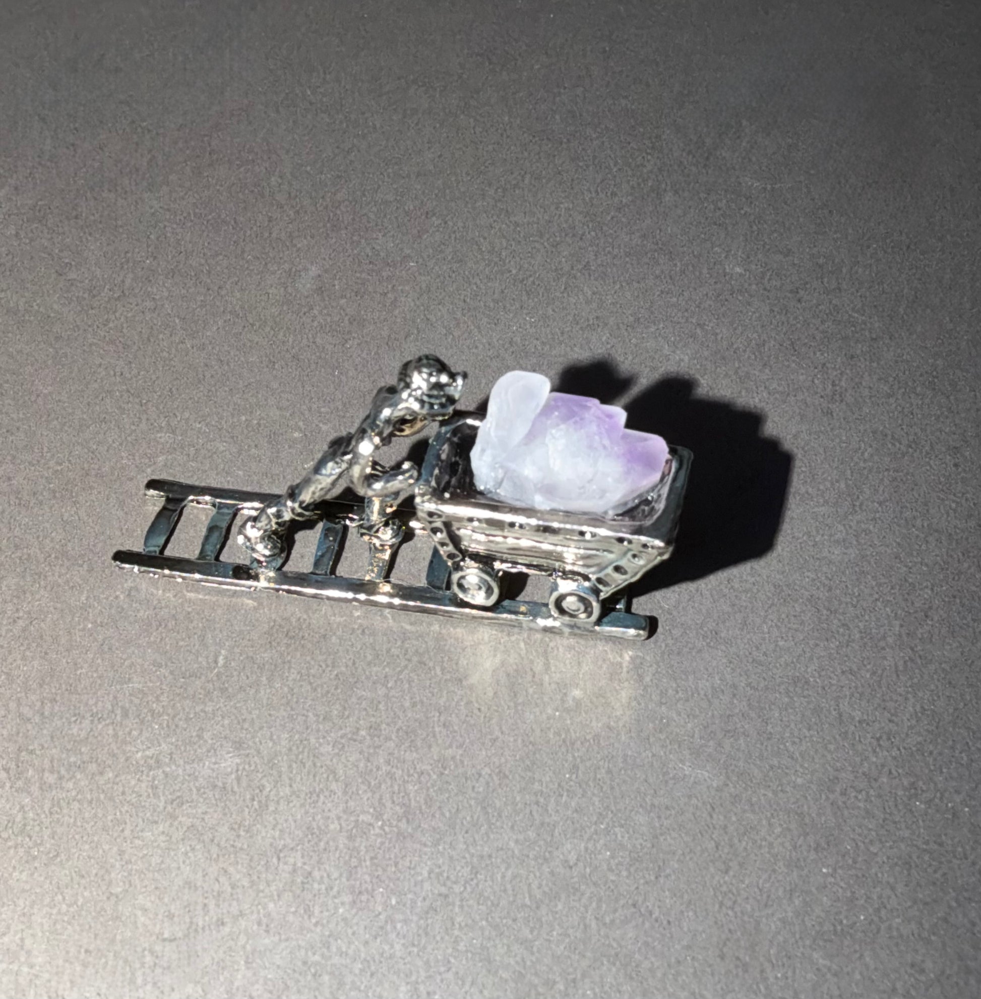 A silver-toned miniature charm depicting a person pushing a sled with a light purple crystal, shown on a gray background.