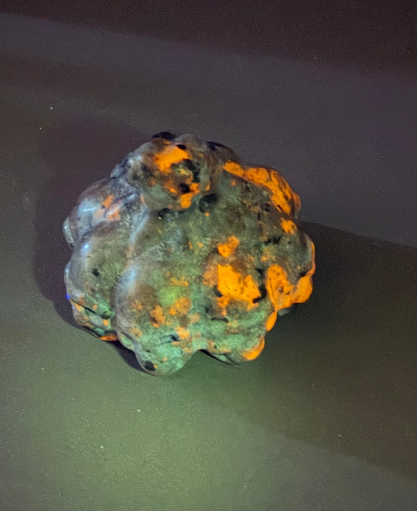 A rough, irregularly shaped specimen with a dark greenish-gray surface and bright orange fluorescent patches, photographed on a dark surface under colored lighting.