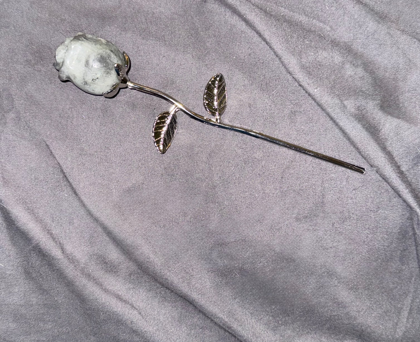 A silver-toned metal rose stem with a textured, stone-like white rose head, resting on a draped gray fabric background.