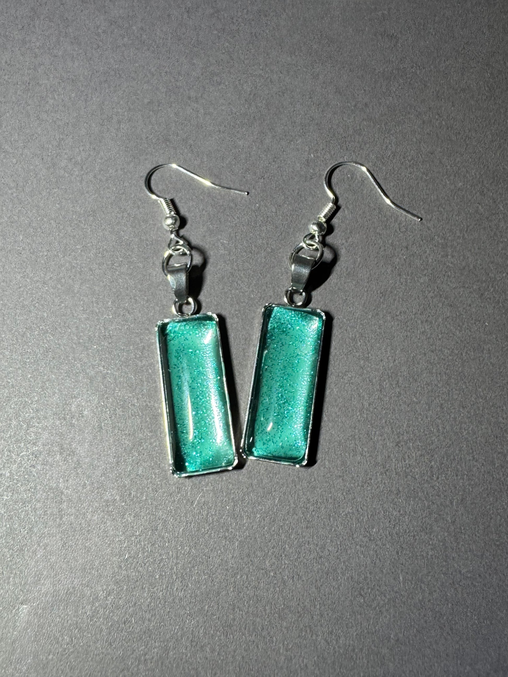 A pair of silver-tone dangle earrings with rectangular green glass pendants, displayed on a textured gray background.