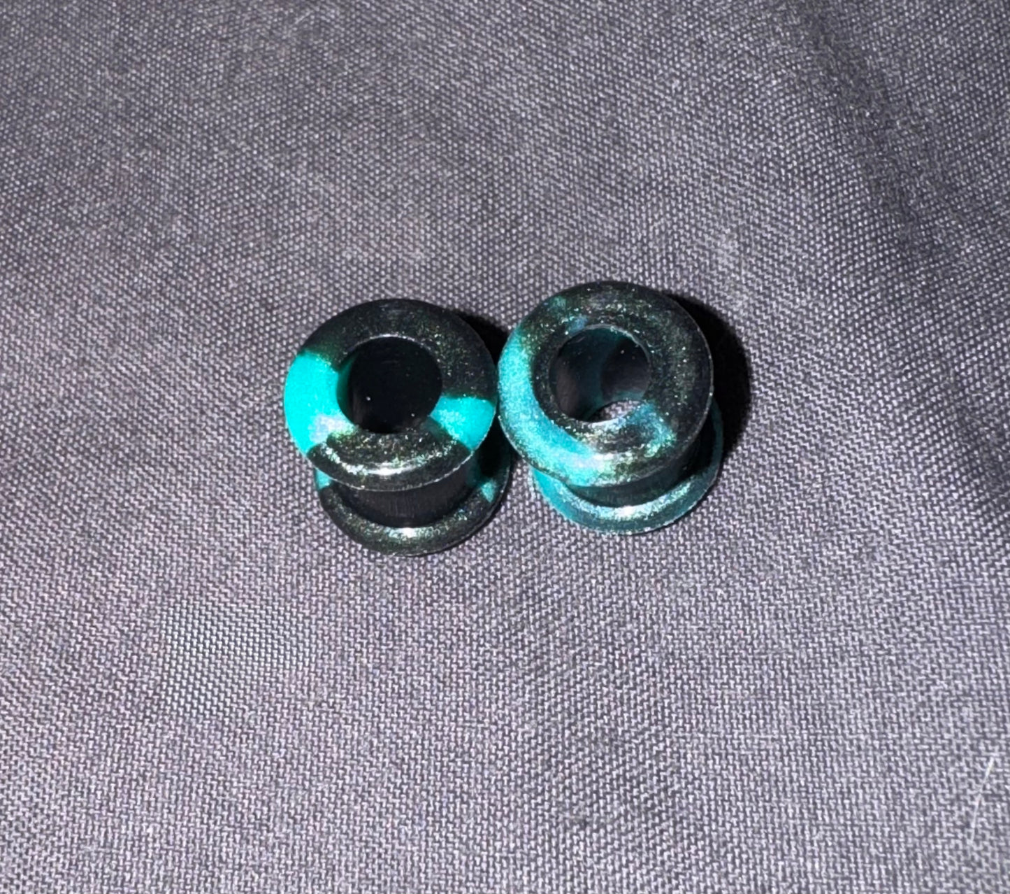 Two small black cylindrical plugs with bright turquoise blue painted accents, placed on a textured gray fabric background.
