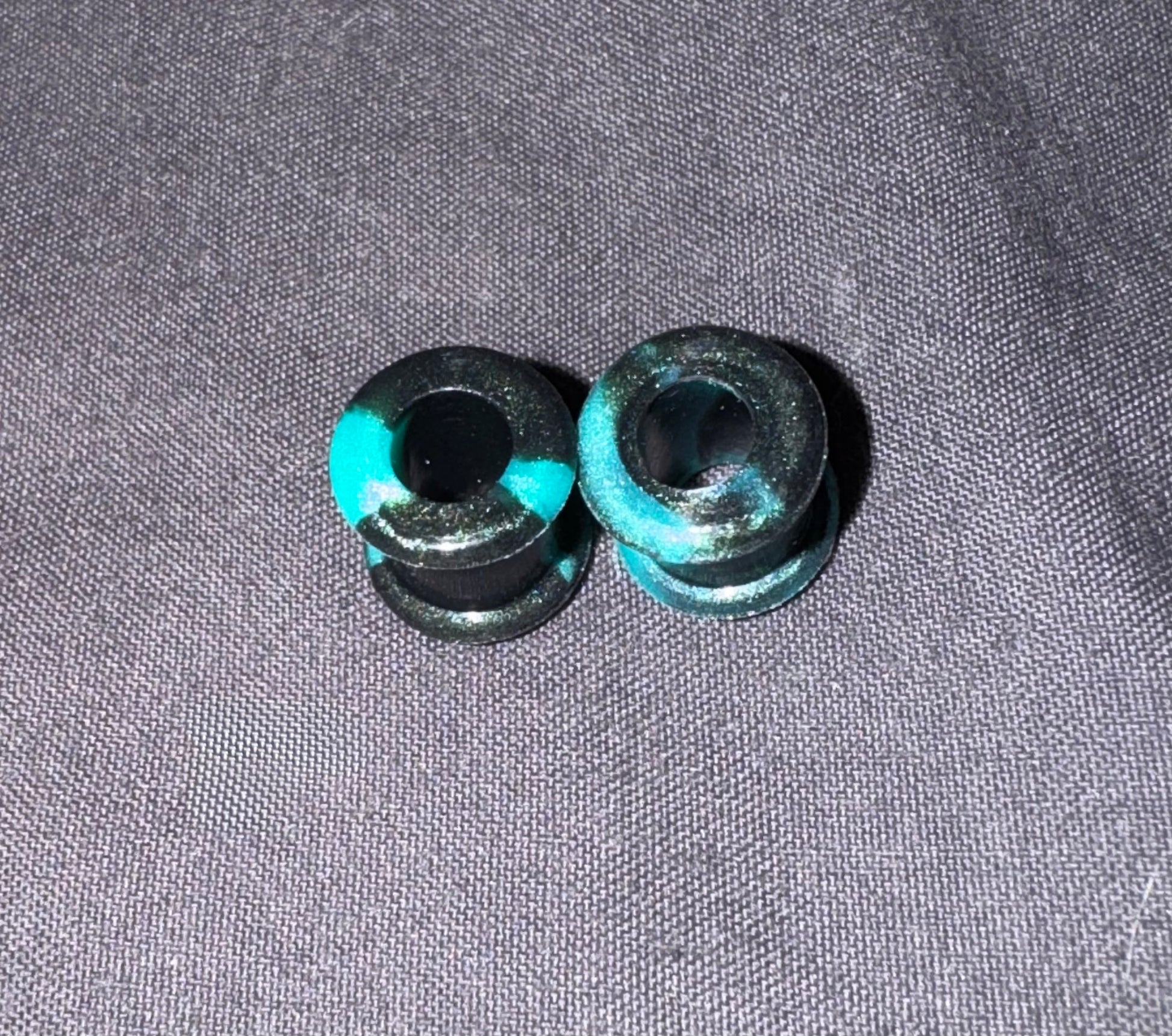 Two small black cylindrical plugs with bright turquoise blue painted accents, placed on a textured gray fabric background.