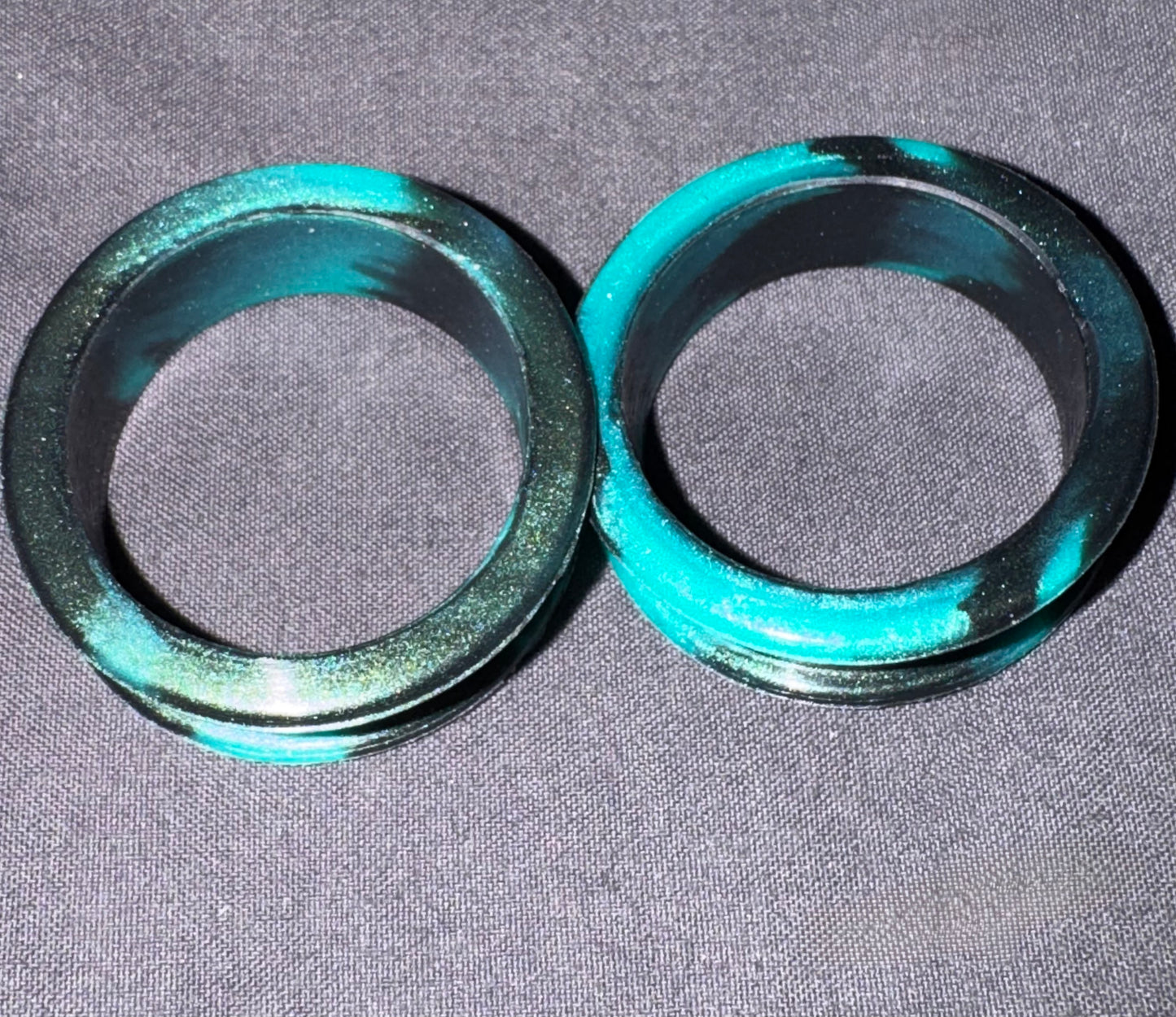 Two circular black rings with a metallic teal-green glittery rim, displayed on a gray textured fabric background.