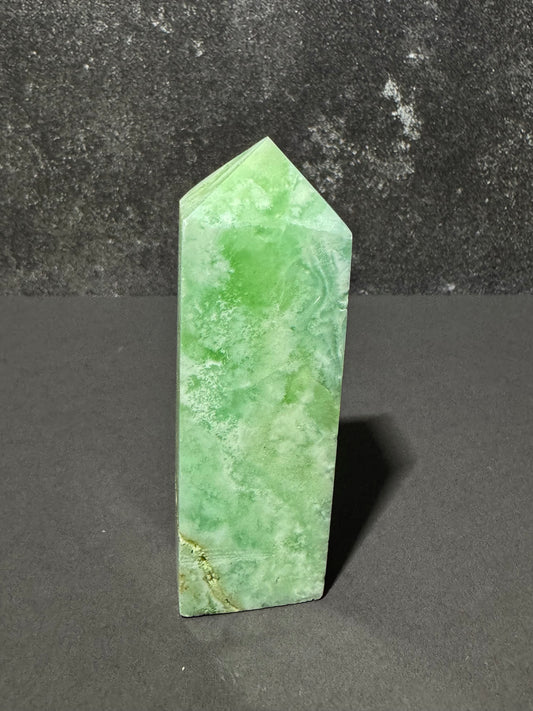 A green stone crystal tower with natural marbled patterns, standing upright on a dark gray surface against a textured dark gray background.