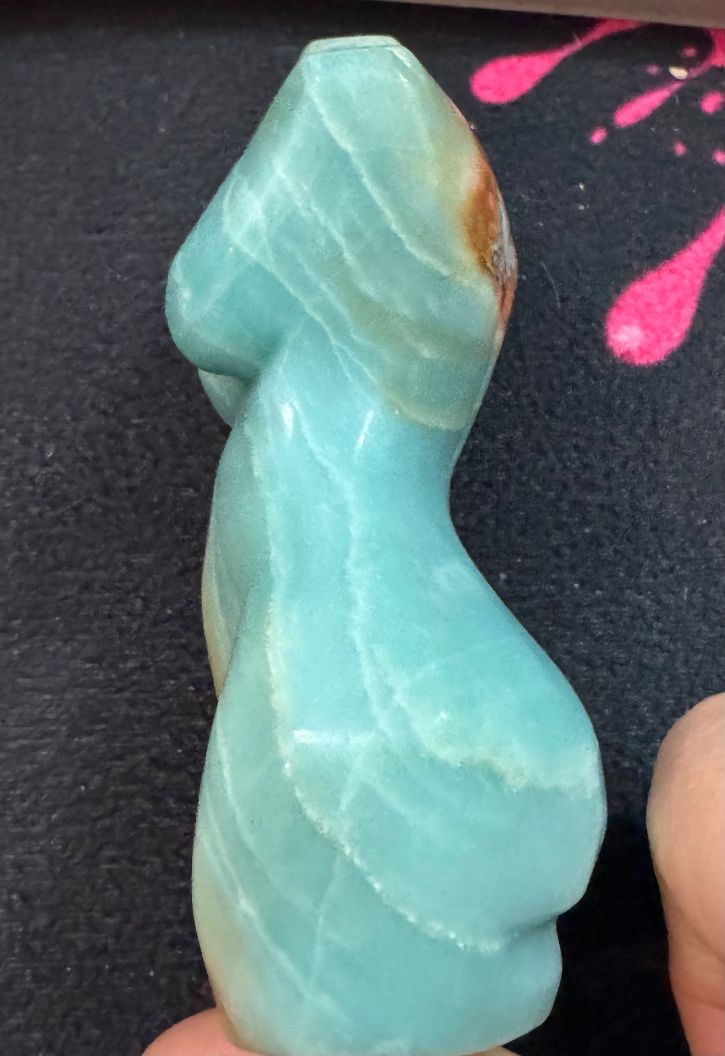 Blue-green stone with a wavy texture held by a hand on a dark surface with pink splashes.