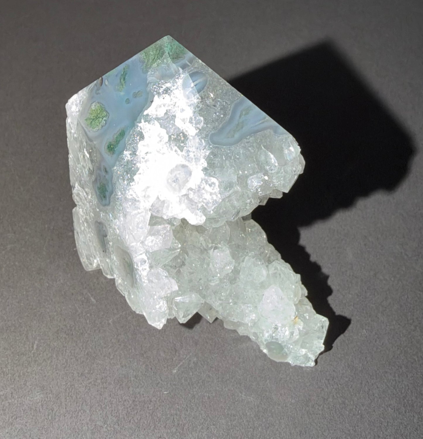 A raw crystal specimen with a pale blue-green and white marbled top section and a rough, translucent white lower section, photographed on a dark gray surface with a sharp shadow.