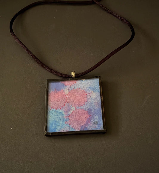 A square pendant necklace with a dark brown cord and a dark metal frame, featuring an abstract watercolor design in pink, blue, and purple. The pendant is displayed on a dark brown background.