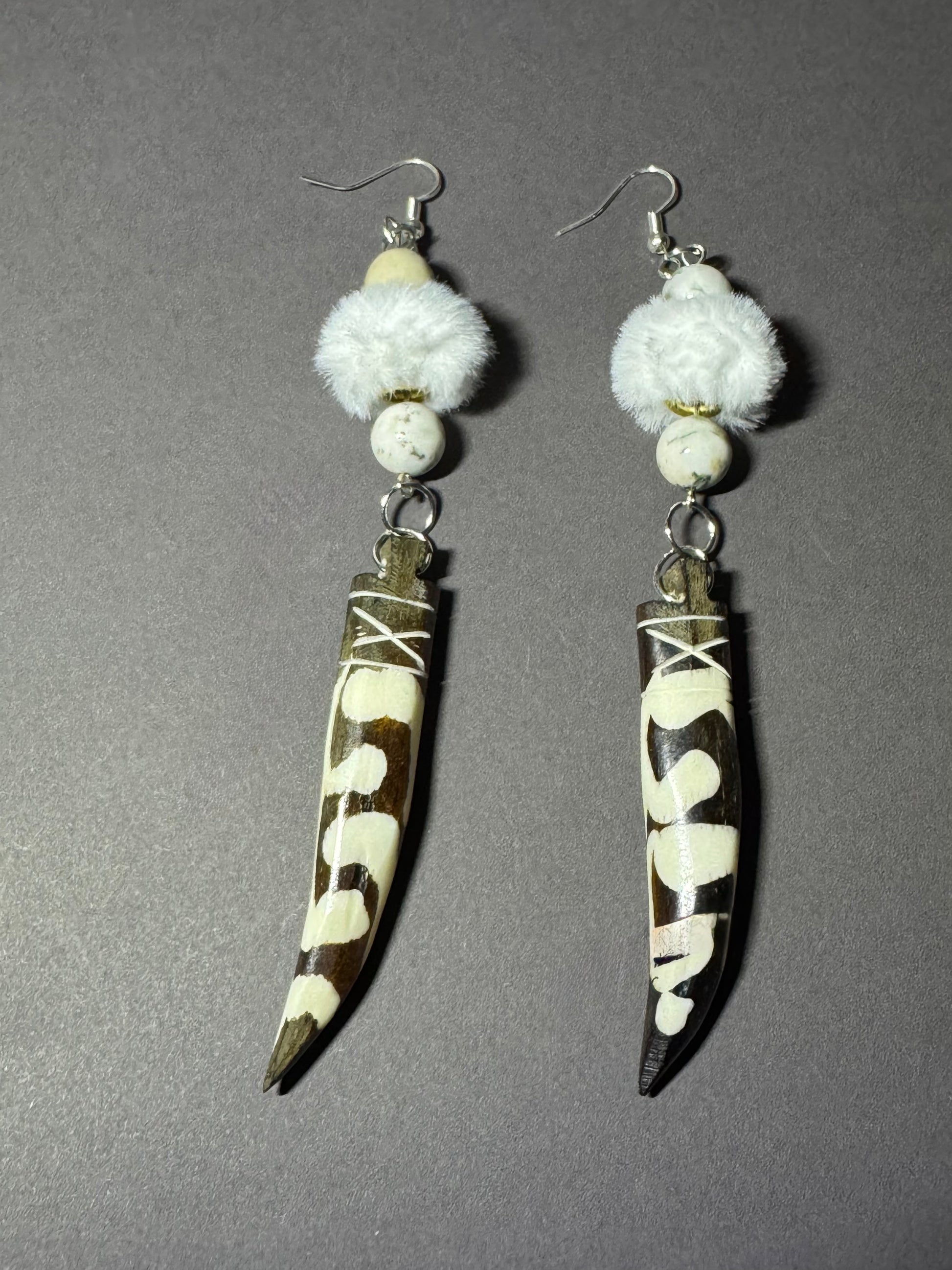 A pair of handmade dangle earrings featuring brown and white cowhide horn blades with white bead details and fluffy white faux fur, displayed on a neutral gray background.